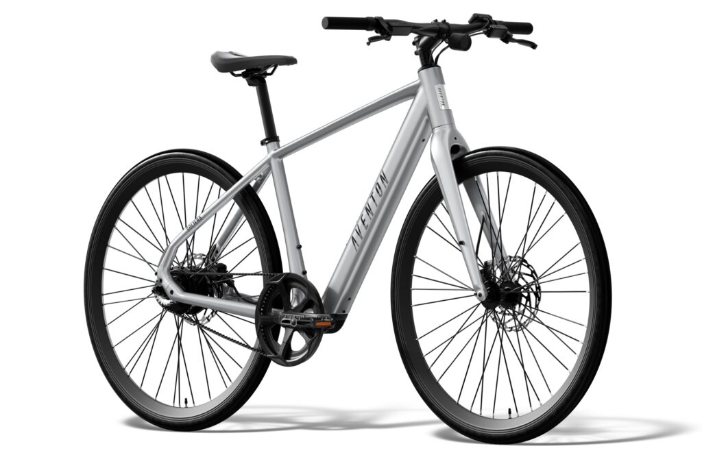 Three-quarter front view of a silver Aventon Soltera 3 ADV commuter e-bike with a belt drive instead of a chain, a flat handlebar and slim tires on a white background.